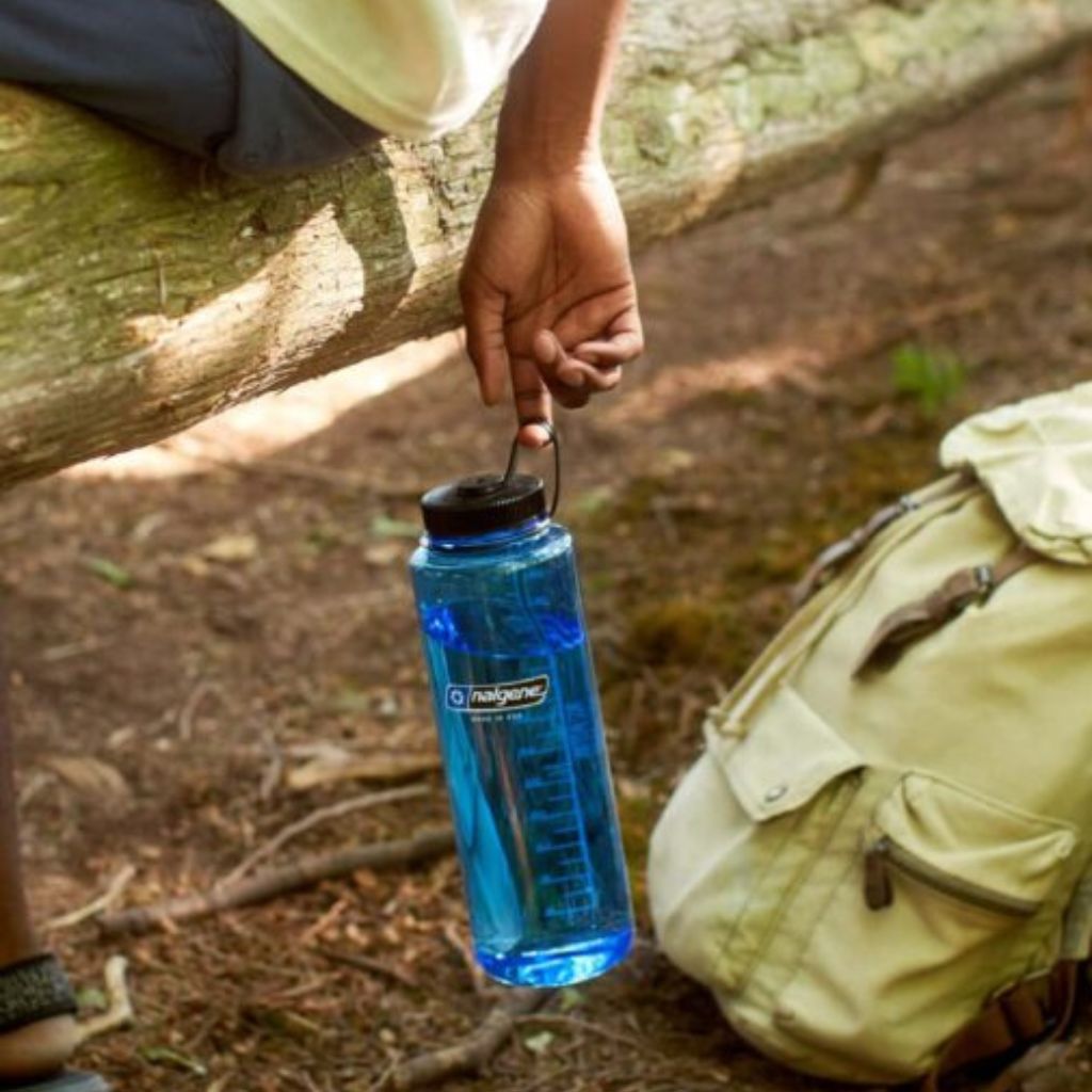 Person holding a blue water bottle with a cap, sitting outdoors next to a backpack.