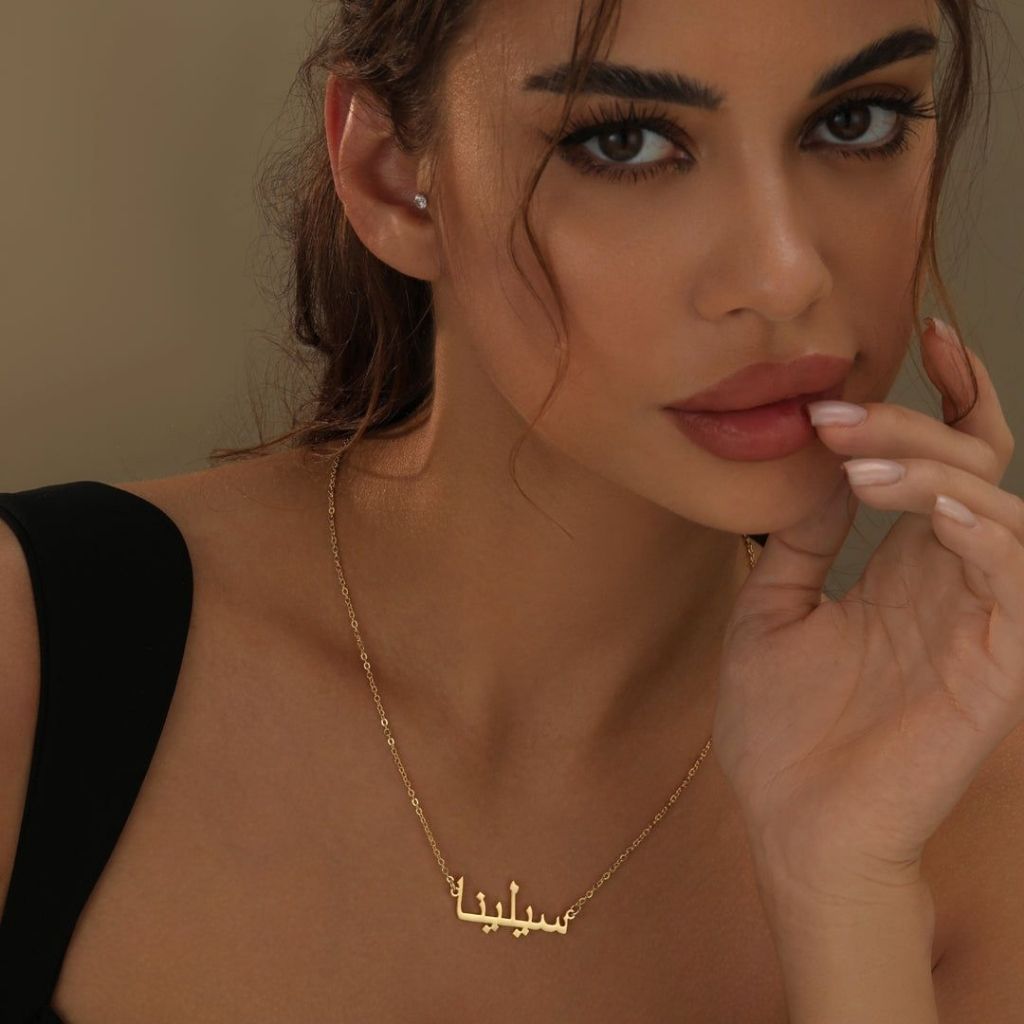 Woman wearing a gold necklace with a nameplate against a neutral background