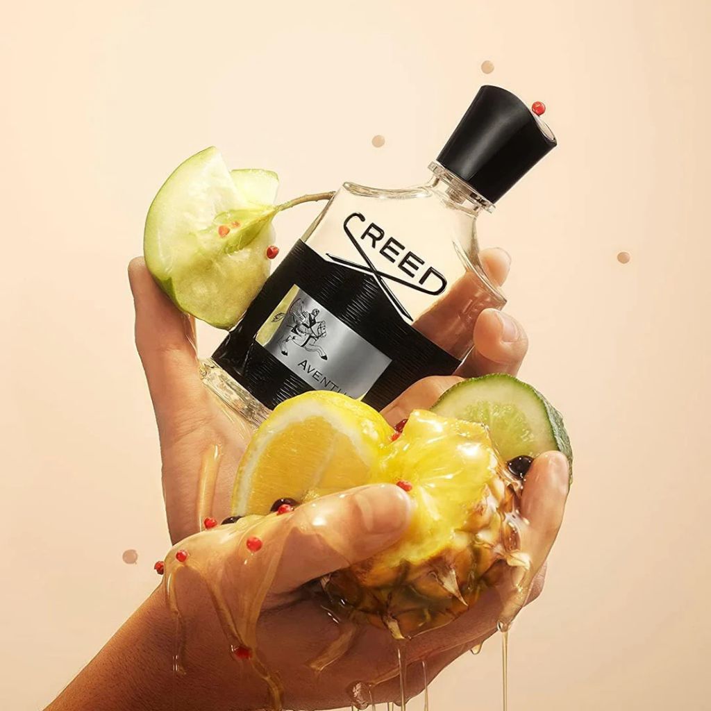 Hand holding a Creed perfume bottle with fruit slices on a beige background