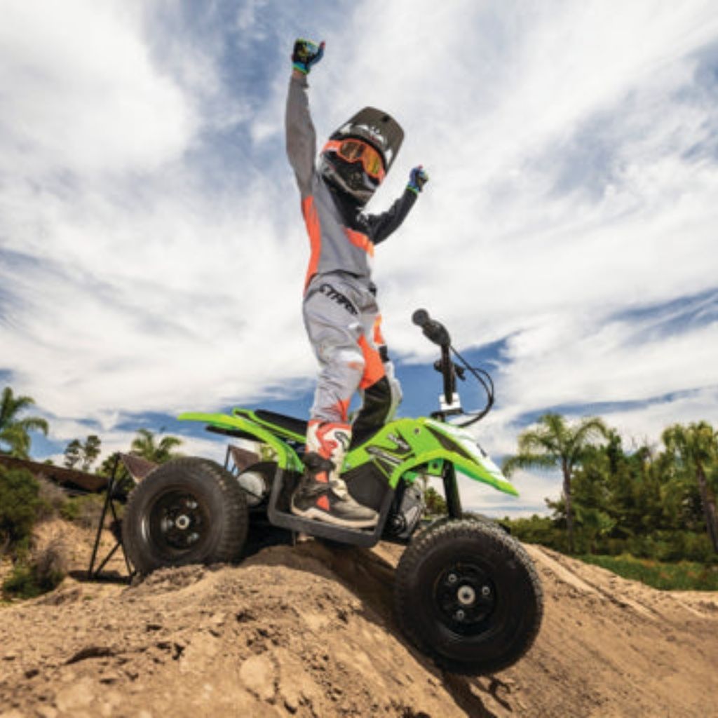 Person on a green ATV with one foot on the ground, celebrating on a dirt hill with trees and sky in the background.