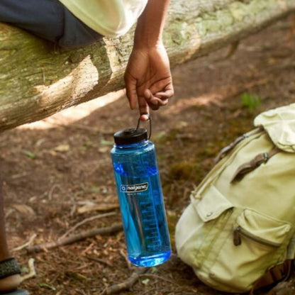 Person holding a blue water bottle with a cap, sitting outdoors next to a backpack.