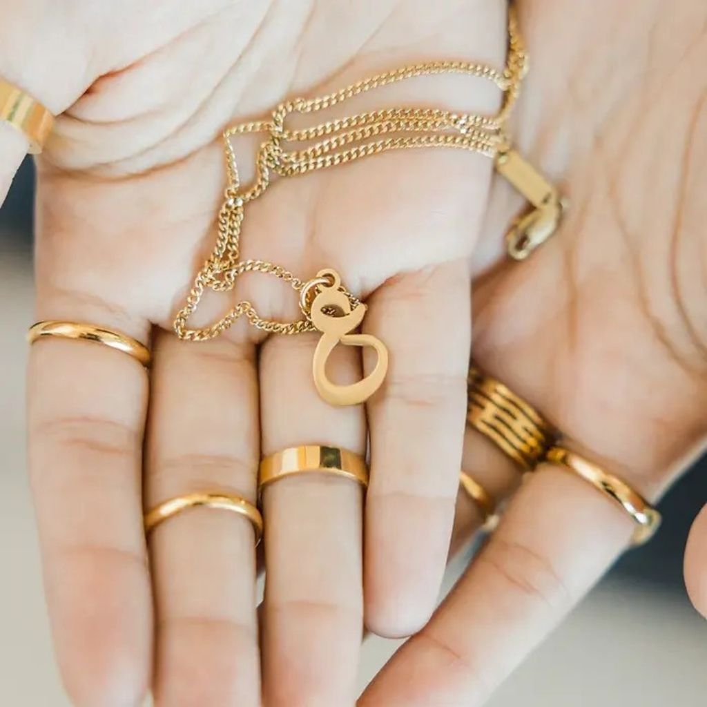 Gold jewelry including rings and a necklace on a hand against a neutral background
