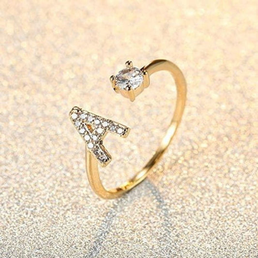 Gold ring with letter 'A' and heart-shaped gem on a glittery beige background
