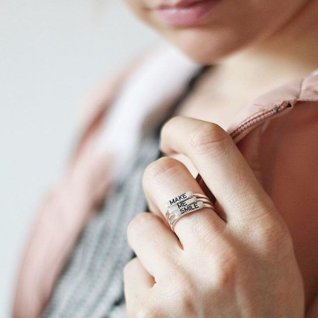 Close-up of a hand wearing a ring with 'MAKE ME SMILE' text on a blurred background