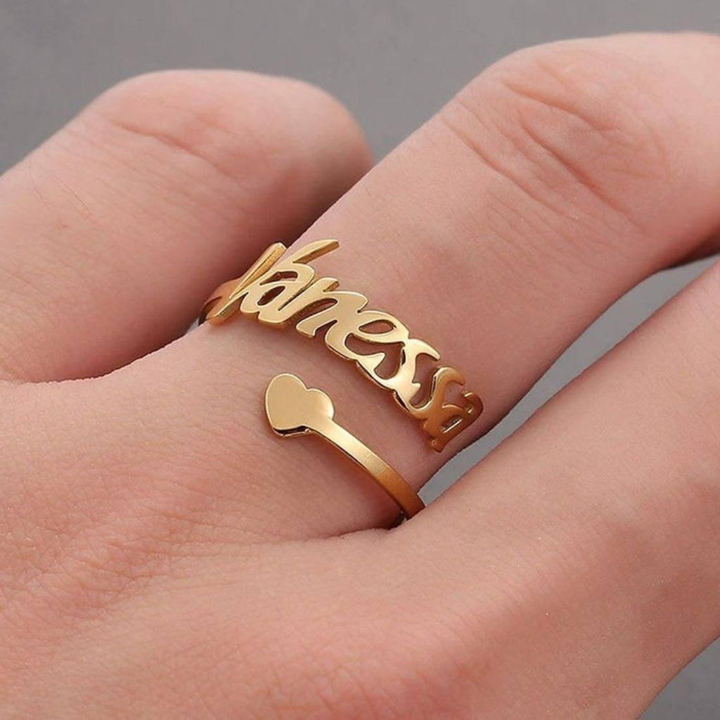 Gold ring with 'fame' and heart design on a hand against a gray background