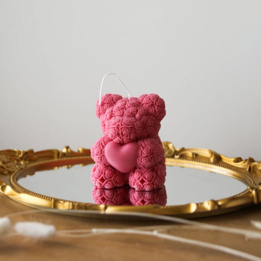 Pink teddy bear-shaped candle on a reflective surface with a gold frame.