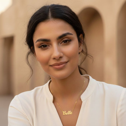 Woman wearing a gold necklace with 'Belle' pendant against a beige architectural background