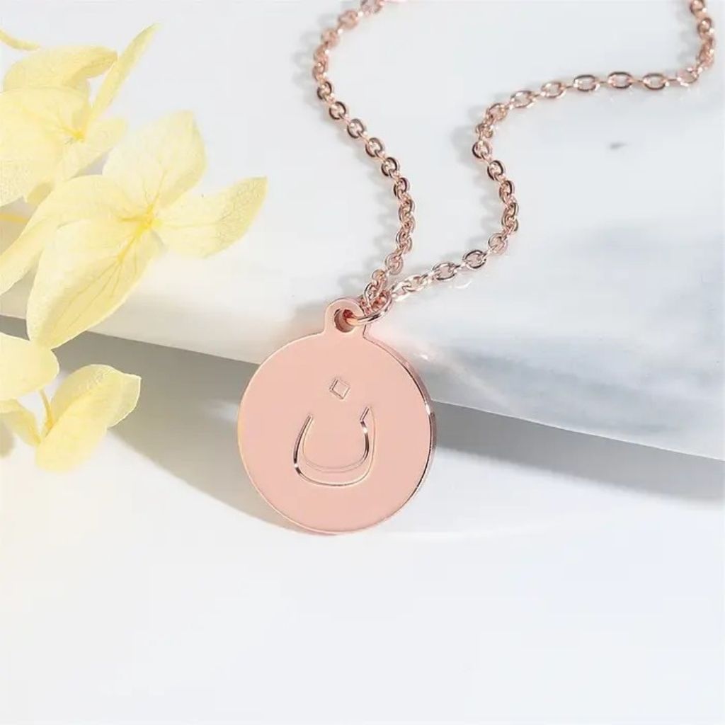 Rose gold necklace with a circular pendant on a white surface with yellow flowers.