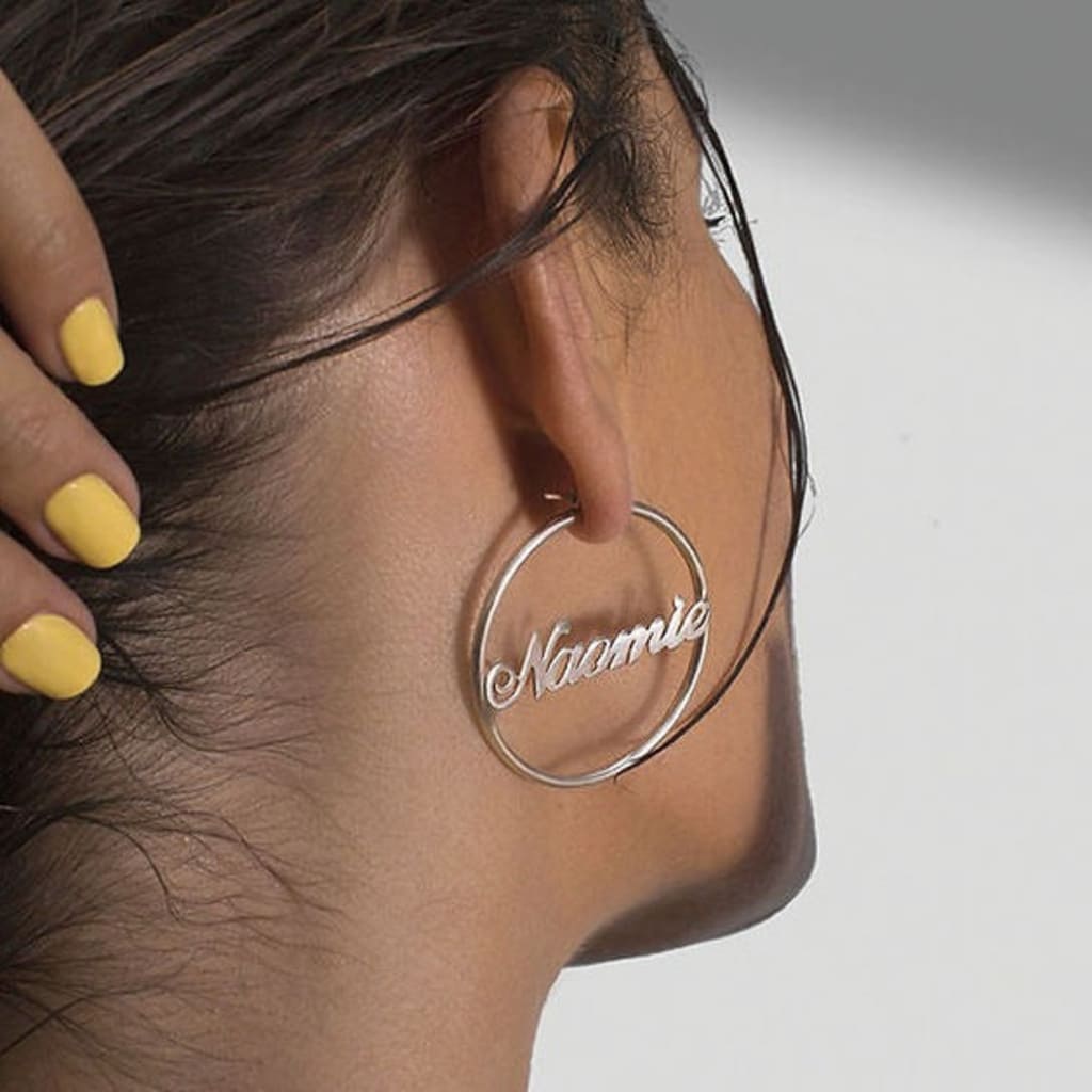 Close-up of an ear wearing a hoop earring with 'Naomie' engraving, against a neutral background.