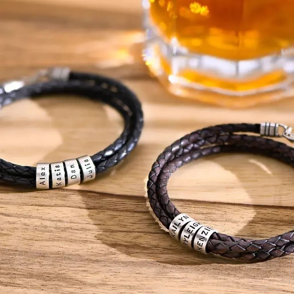 Two braided leather bracelets with metal name beads on a wooden surface, with a glass of amber liquid in the background.