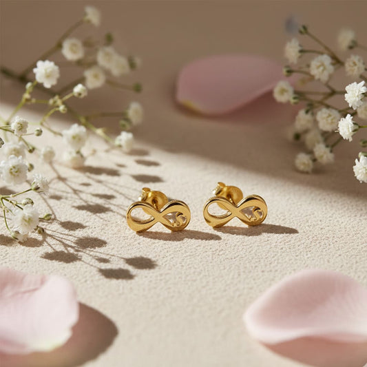 Gold infinity symbol earrings on a beige surface with white flowers and pink petals.