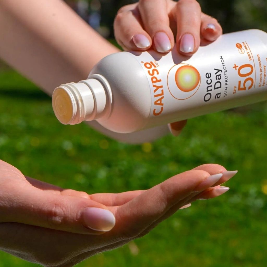 Person applying Calypso 'Once a Day' sunscreen spray to their hand with a green outdoor background.