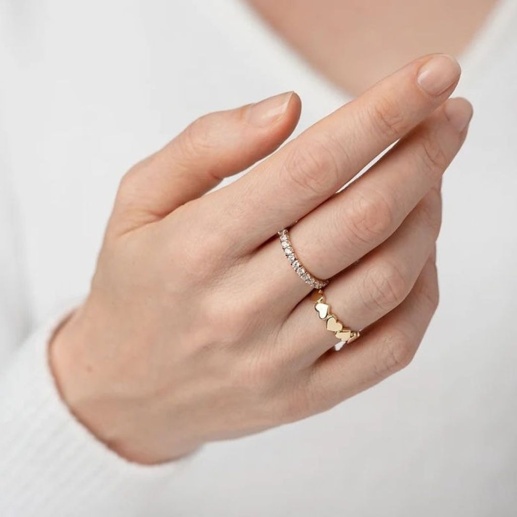 Hand wearing two gold rings with heart designs on a light background