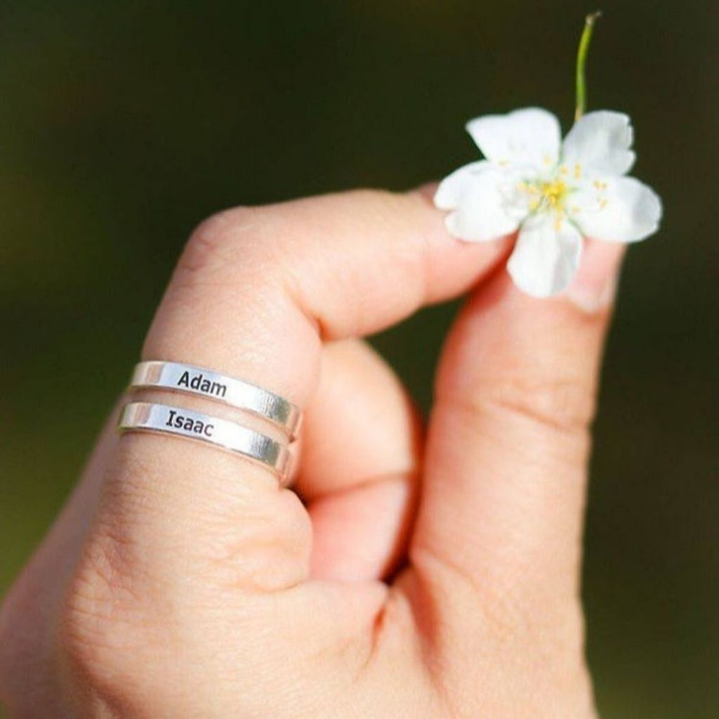 Hand wearing two silver rings with engraved names, holding a small white flower.