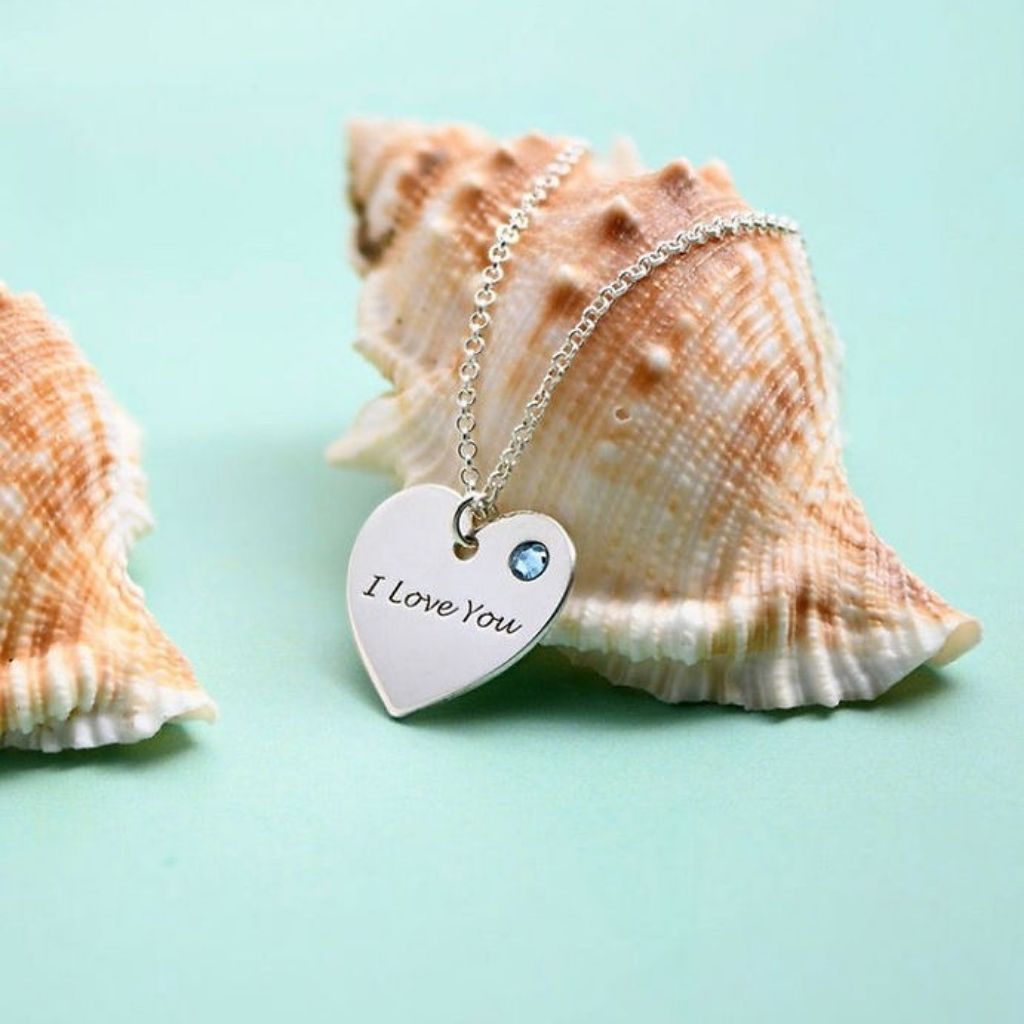 Heart-shaped pendant with 'I Love You' engraving on a chain, placed on a shell against a light blue background.