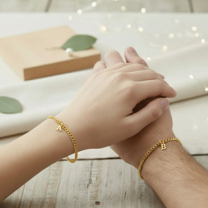 Two hands clasped together wearing gold bracelets with letters on a wooden surface.