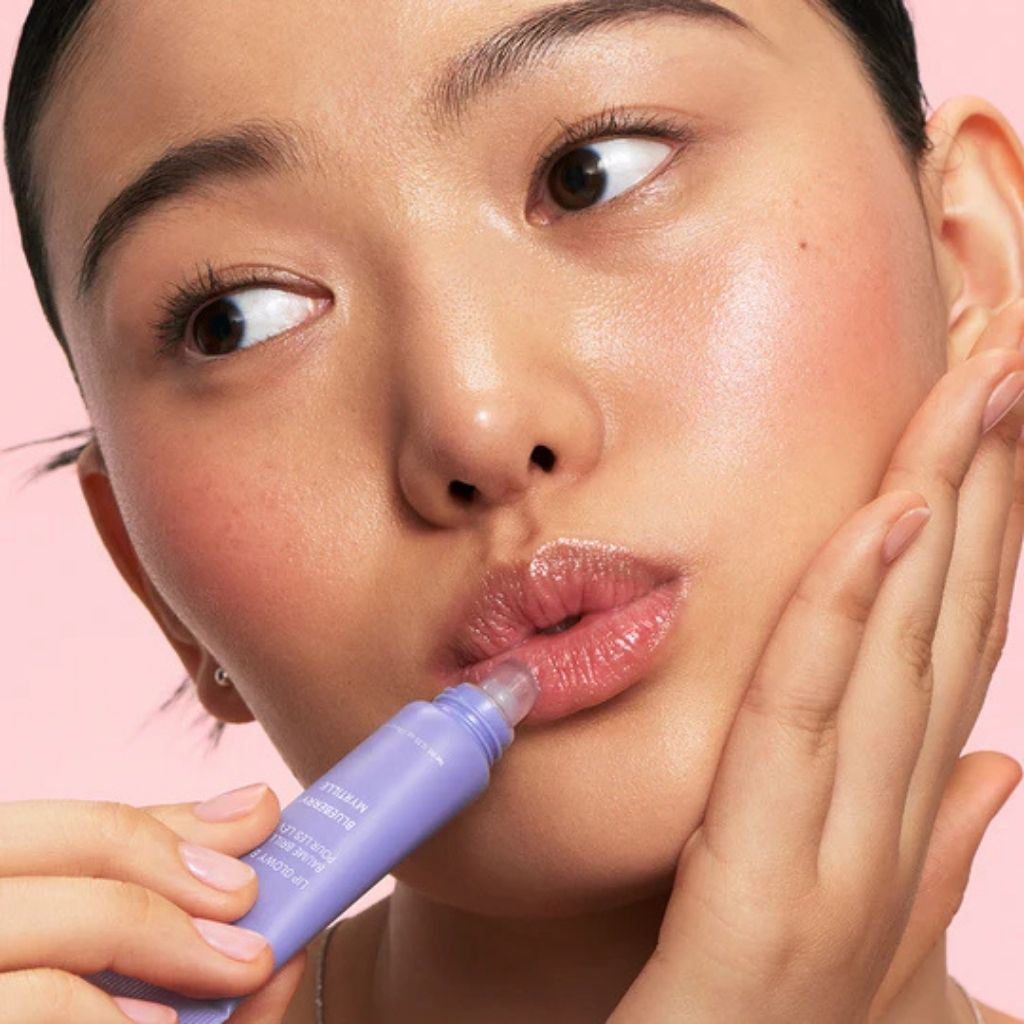 Woman applying a purple lip product with a pink background