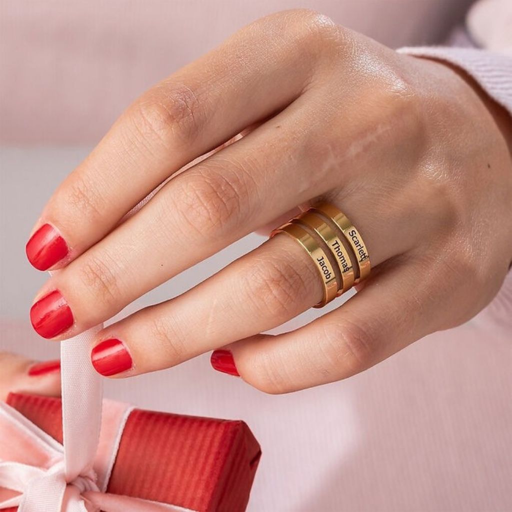 Hand with red nail polish wearing gold rings on a light pink background