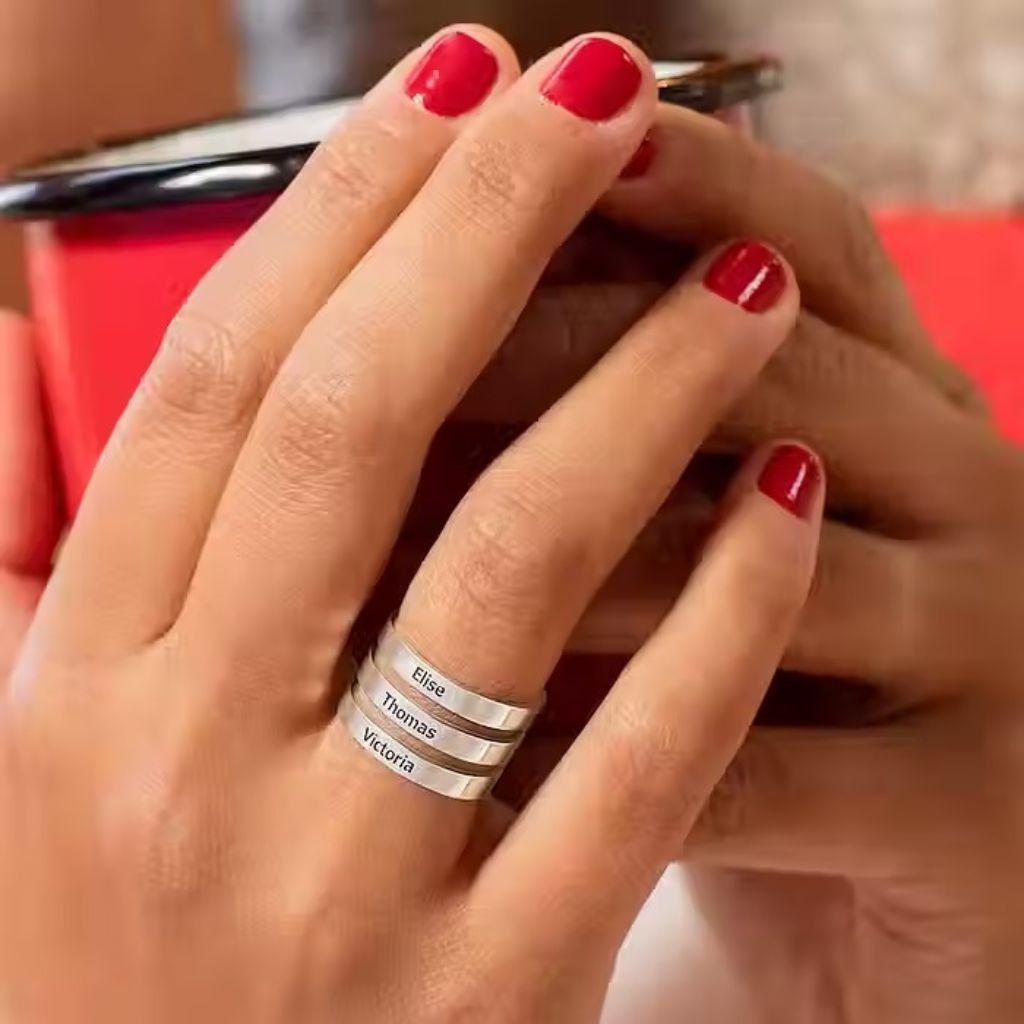 Hand wearing a silver ring with engraved names, red nail polish on other fingers.