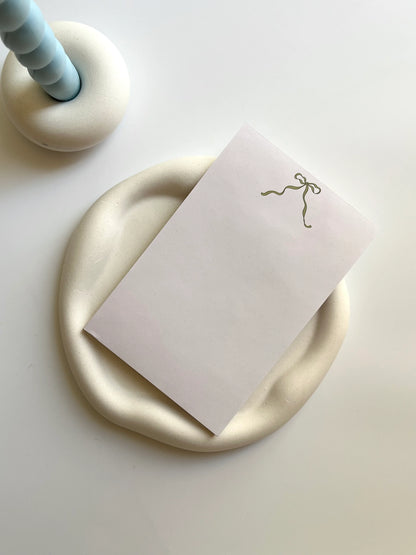 White card with a gold bow on a gold circular object, possibly a coaster, on a light surface.