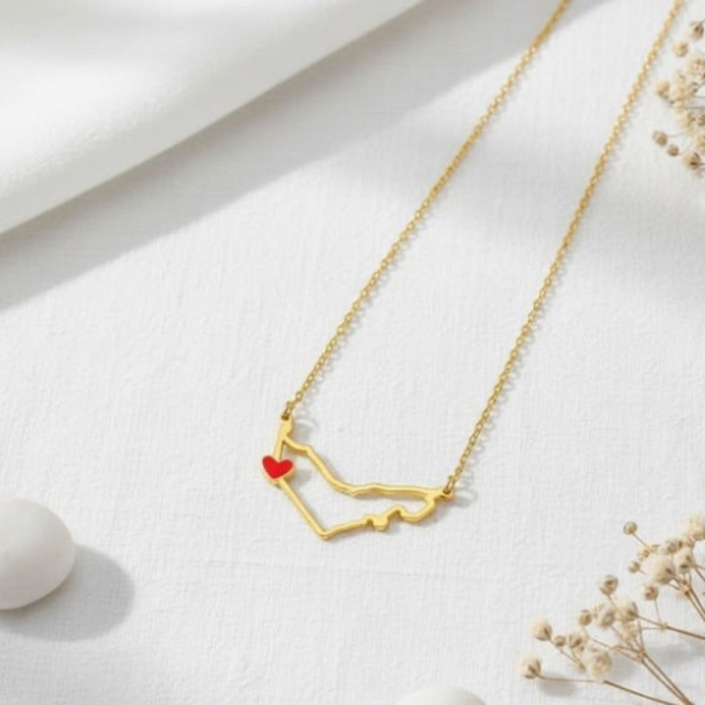 Gold necklace with a red heart pendant on a white surface with dried flowers.