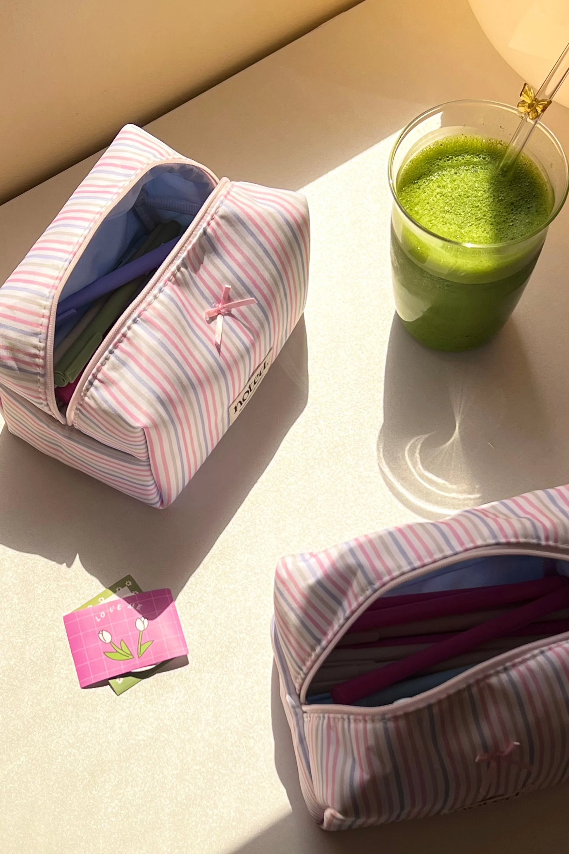 Striped makeup bags on a light surface with a glass of green juice.