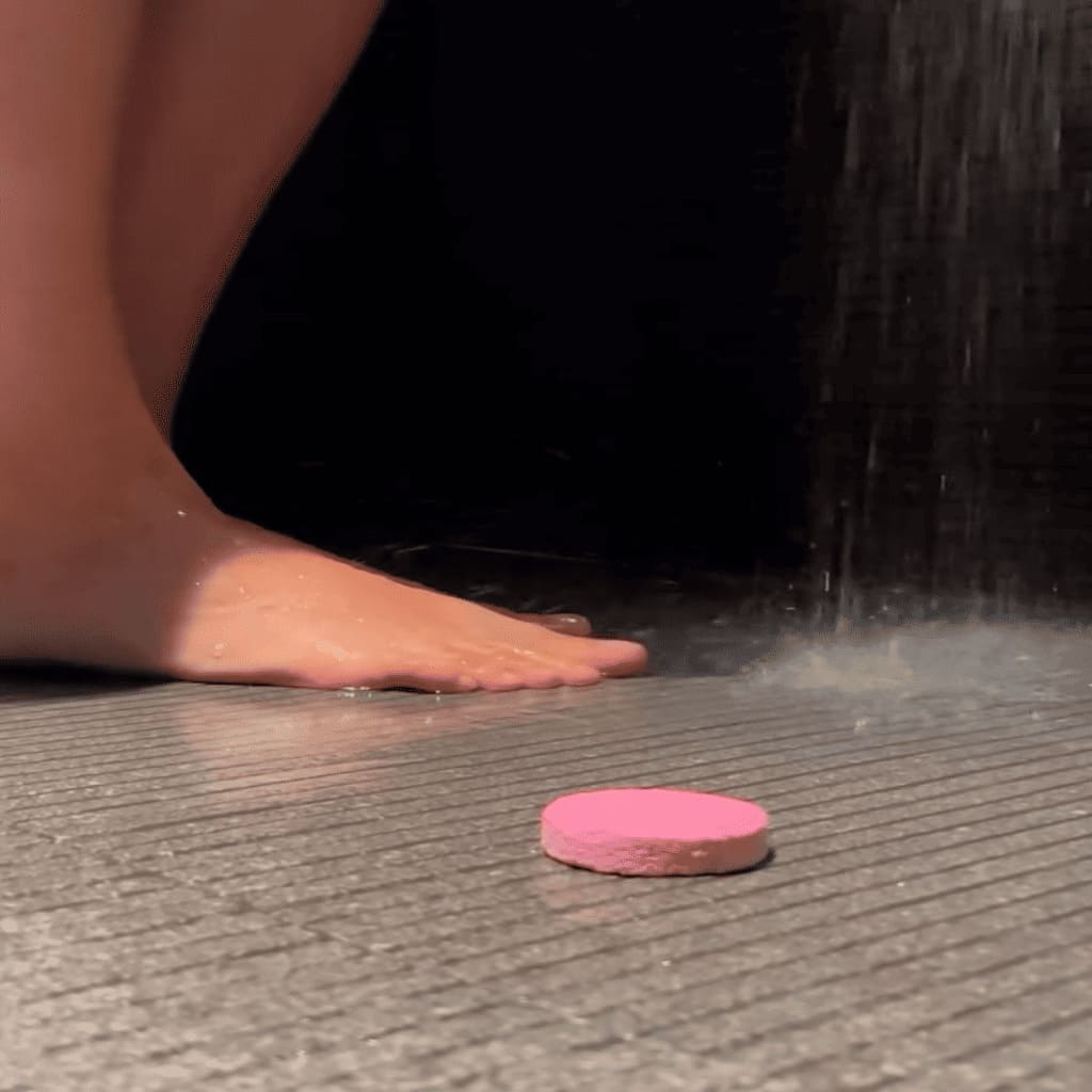 Pink bath bomb on a tiled floor with water droplets and a shower in the background