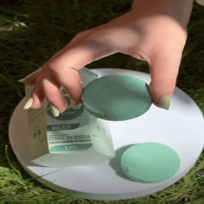 Hand holding a green bath bomb over a white plate with more bath bombs on a grassy background