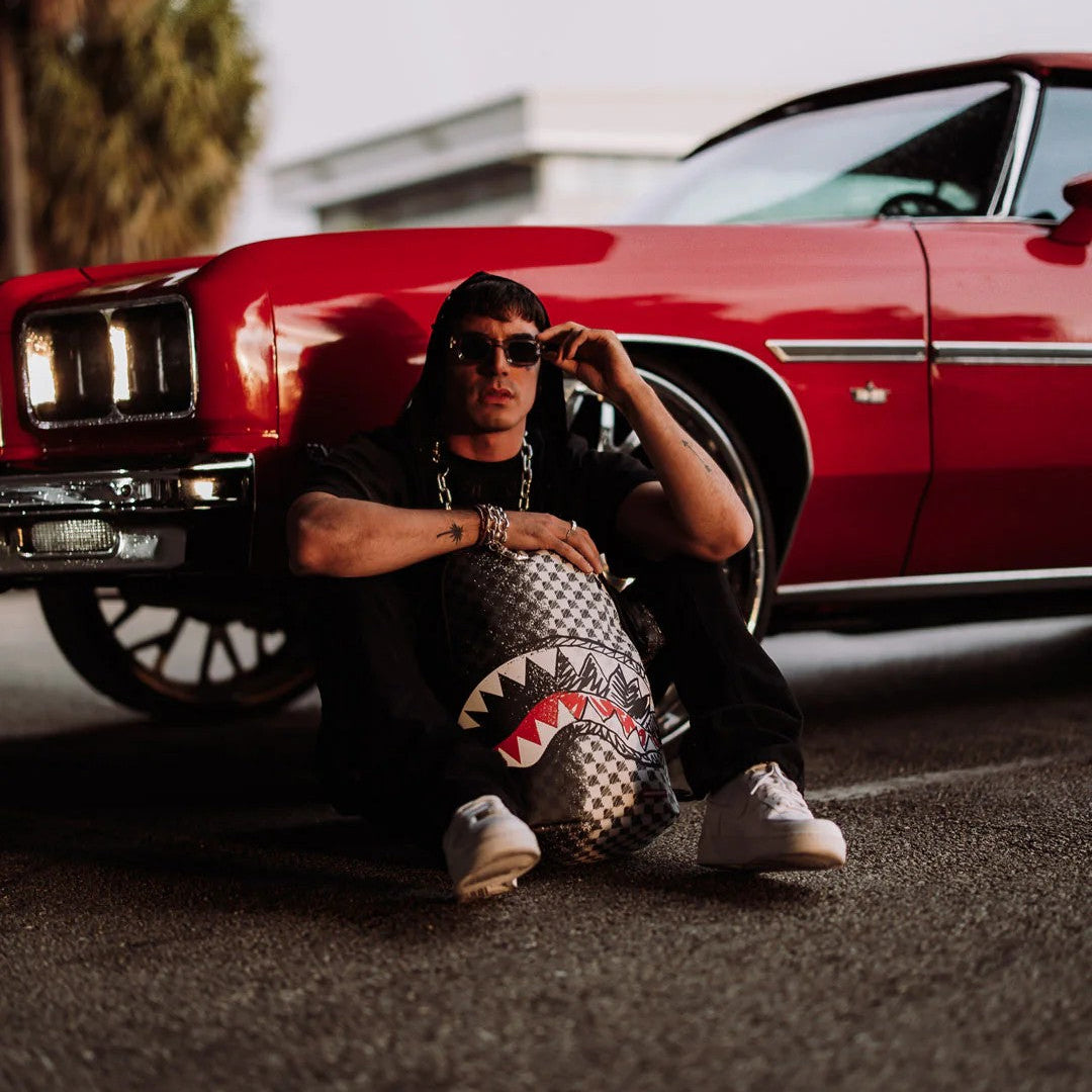 Person sitting on the ground next to a red classic car, holding a patterned bag.
